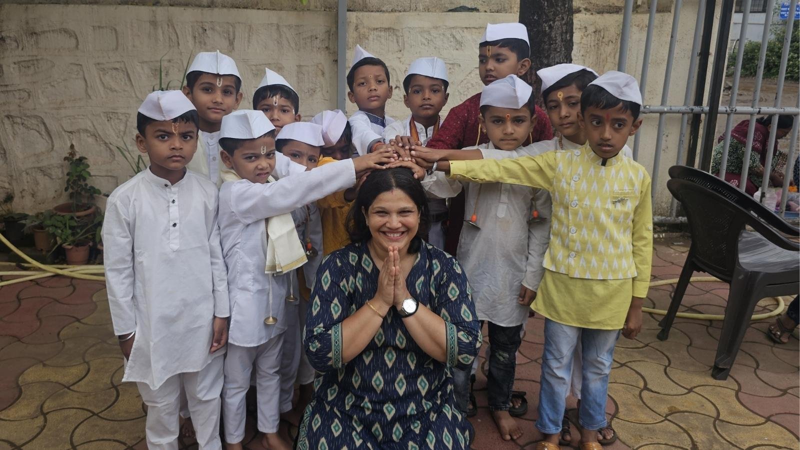 Aarati kneeling and smiling with her hands folded in namaste, surrounded by a group of young children in traditional white clothing who are affectionately placing their hands on her head.